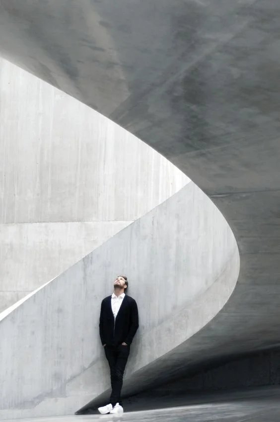 Persona de pie en una estructura de concreto moderna y minimalista, observando hacia arriba en un entorno arquitectónico abstracto.